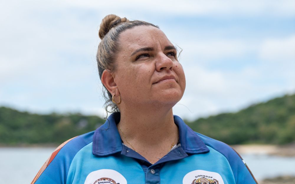 Woman stands on beach looking out into the distance in blue collared shirt. 