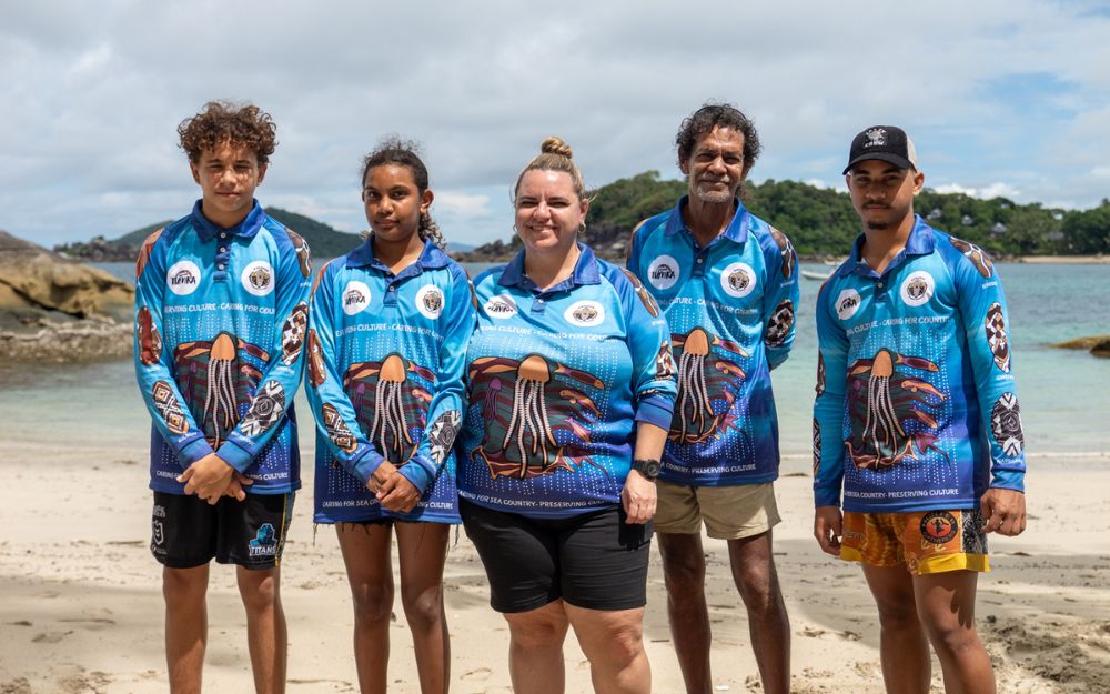 Five people stand on beach of Australian First Nations descent. 