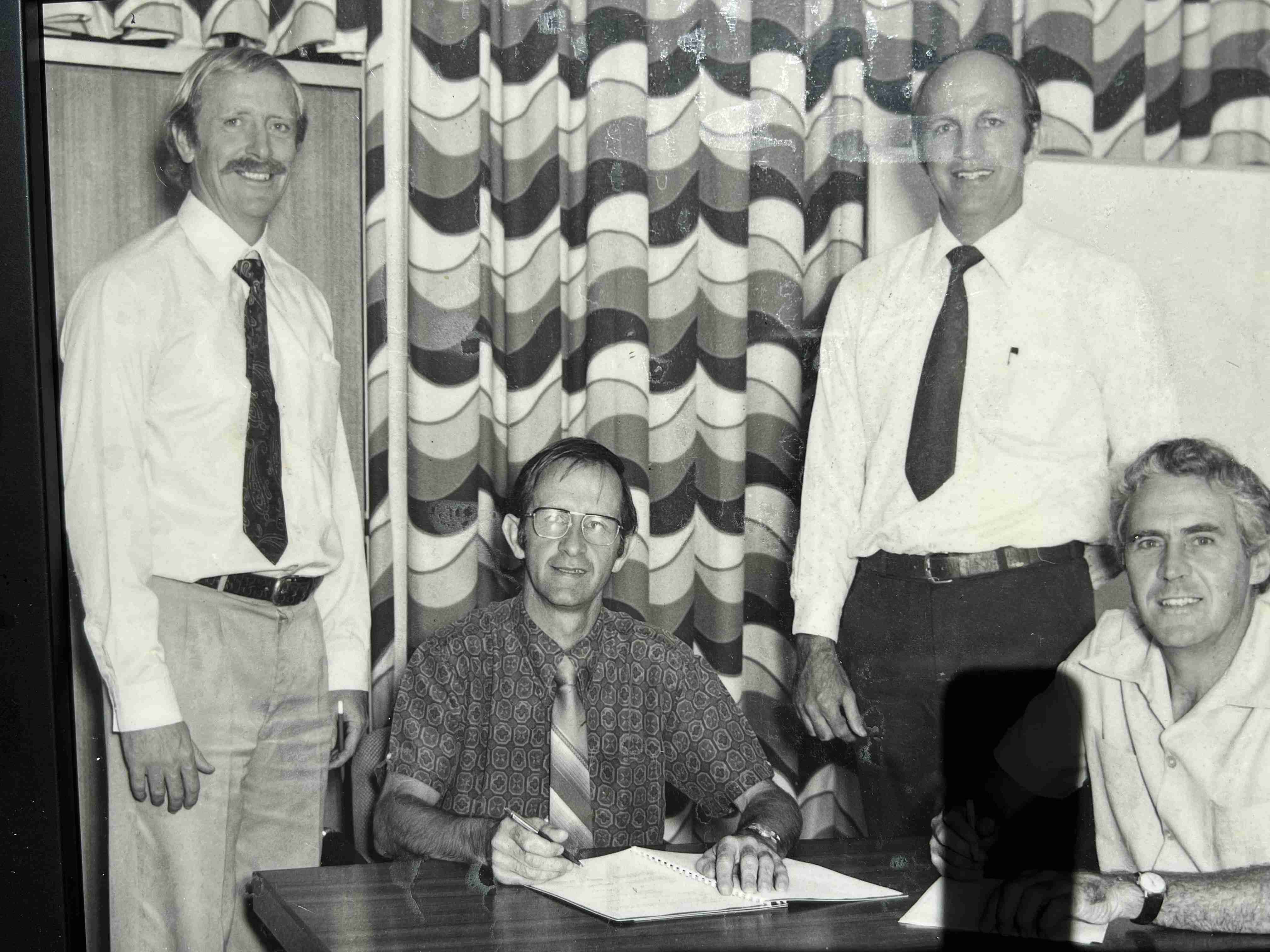 Black and white photo of four men posing for photo while document is signed