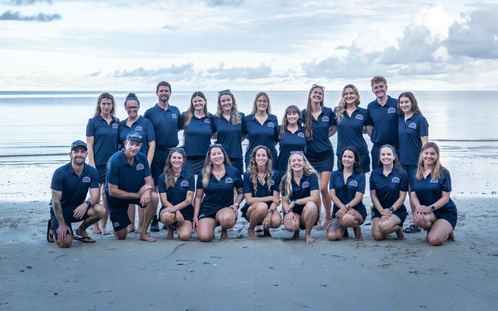 A group of people in navy polo shirts poses for a photo on a beach.