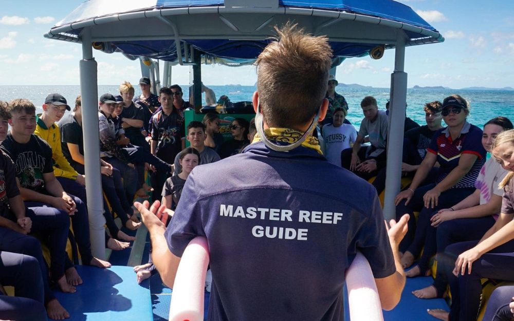 Man with his back to the camera stands on boat in front of school children to deliver a presentation.