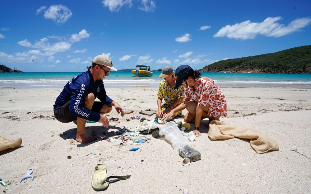 Three people crouch on beach to pick up litter with turquoise water in background.