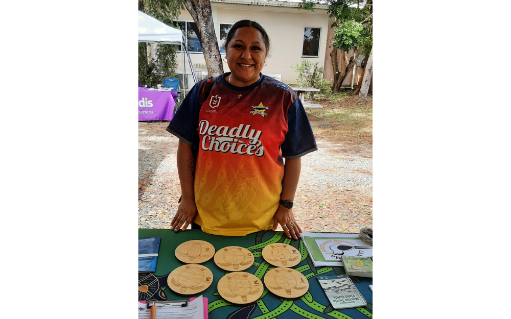 A Lama Lama female ranger stands beside a table containing six wooden turtle discs.