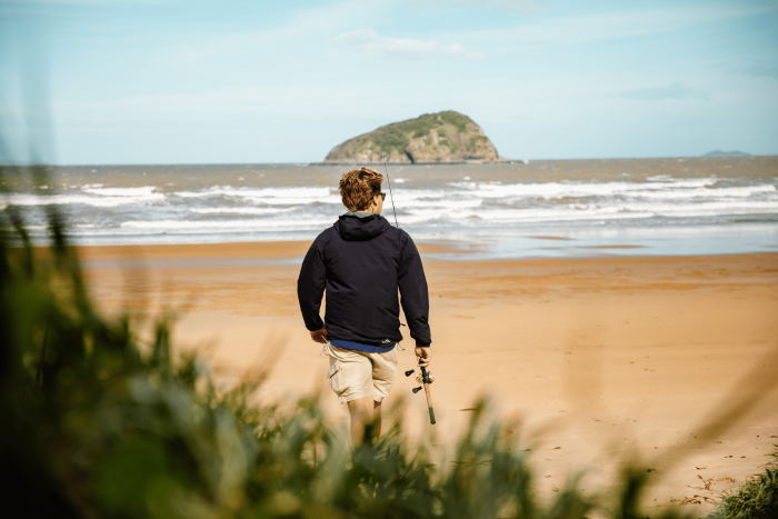 Man seen from behind, walking towards the beach with a fishing rod in hand