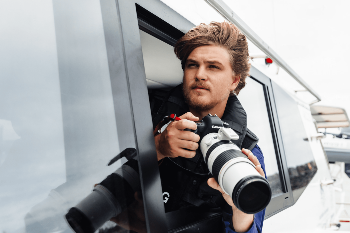 Man leans out of a boat window, holding a Canon camera with a large telephoto lens