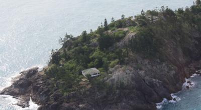 An island with pine trees hosts one small cottage and a helipad. 