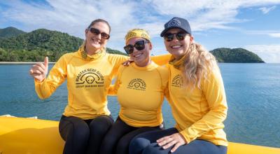Three women in yellow shirts sit on the edge of a boat.