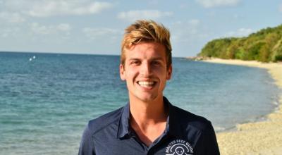 A man stands on a beach with thick vegetation in the background.