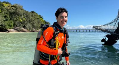 A woman commercial diver in a bright orange rash shirt stands in waist deep ocean water against the backdrop of a tropical island.