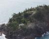 An island with pine trees hosts one small cottage and a helipad. 
