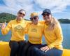 Three women in yellow shirts sit on the edge of a boat.