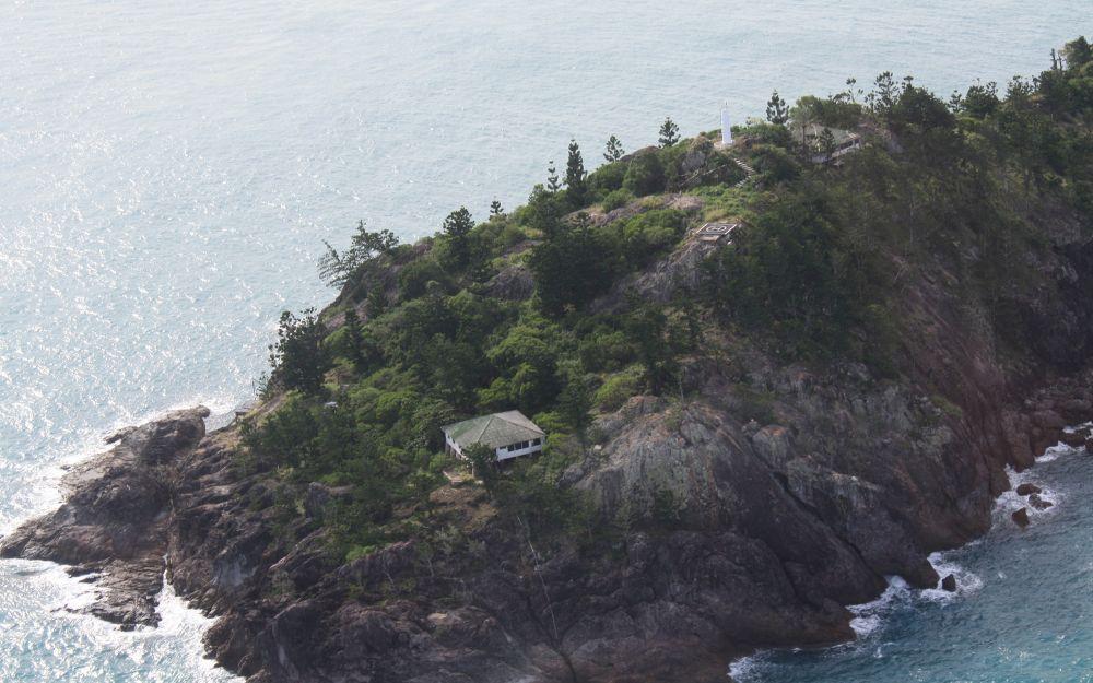 An island with pine trees hosts one small cottage and a helipad. 