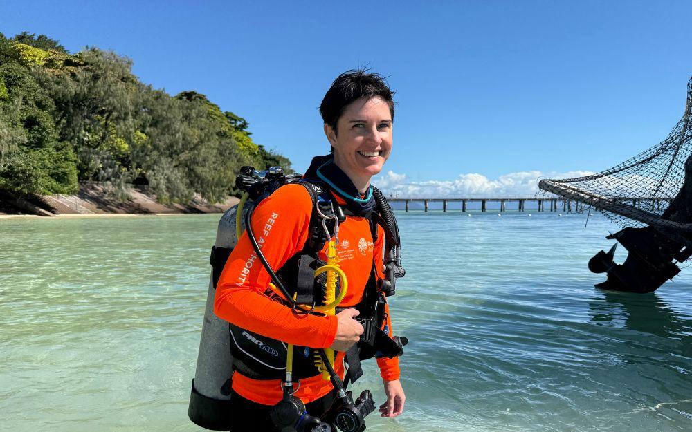 A woman commercial diver in a bright orange rash shirt stands in waist deep ocean water against the backdrop of a tropical island.
