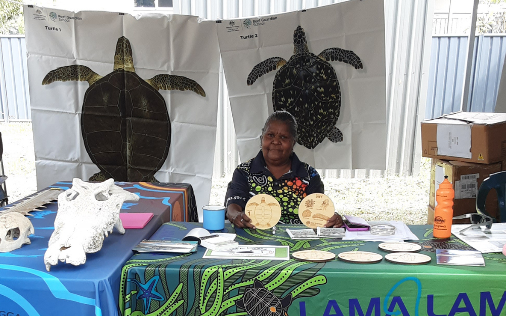 An Aboriginal ranger sits at a community event stall holding wooden discs with turtles on them with turtle posters behind her.