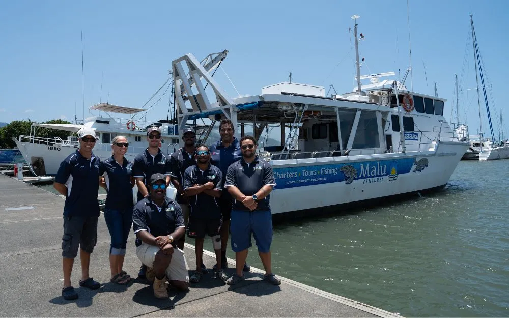 A group of people stand on a jetty in a marina with a commercial boat in the background